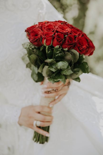 A close-up of a bride holding a bouquet of vibrant red roses with dark green foliage, wrapped in a beige ribbon. The bouquet is arranged in a round, compact style, featuring fully bloomed roses with v