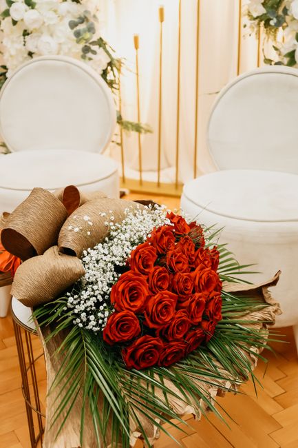 A bouquet arrangement of vibrant red roses, complemented by delicate white baby's breath and lush green palm leaves, is placed on a table in a well-lit room. The bouquet is wrapped in rustic brown pap