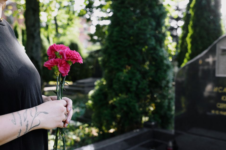 A person holding a bouquet of vibrant pink carnations with ruffled petals and green stems, standing outdoors in a lush, sunlit setting. The individual is wearing a black top and has a visible tattoo o