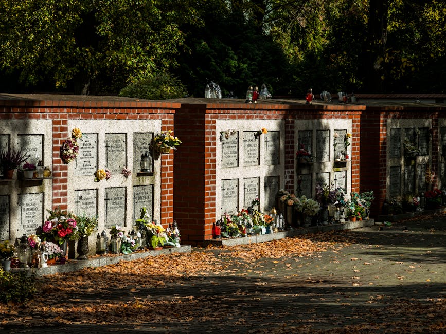 A row of memorial plaques set into a brick and stone wall at a cemetery, each decorated with fresh floral arrangements. The flowers include a variety of colours such as pinks, yellows, purples, and wh