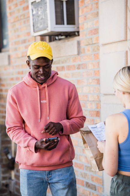 A young delivery person wearing a yellow cap and a pink hoodie, smiling as they hand over a floral gift to a woman with blonde hair wearing a blue sleeveless top. The floral arrangement is inside a br