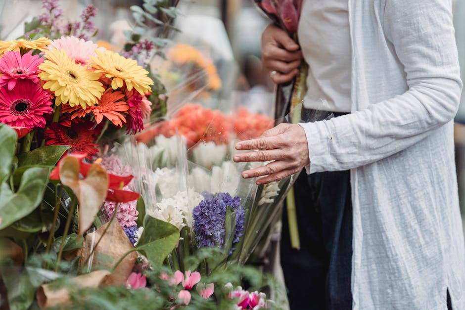 A person wearing a light grey cardigan over a white shirt is selecting a floral arrangement at a flower market, surrounded by a vibrant display of fresh flowers including pink, yellow, orange, purple,