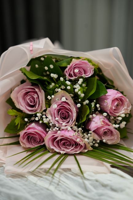 A bouquet of fresh pink roses with delicate petals, arranged with lush green leaves and small white baby's breath flowers. The bouquet is wrapped in elegant white tissue paper with a soft pink ribbon